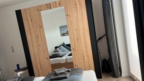 Un dormitorio con cama, espejo, ventana con cortinas y suelo de madera.