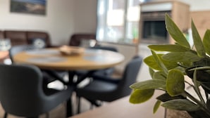 Un área de comedor con una mesa redonda y sillas, una cocina al fondo y una planta en maceta al frente.