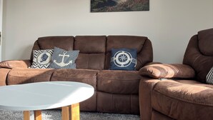 Un sofá de cuero marrón con dos almohadones decorativos, una mesa de centro blanca y un cuadro de paisaje en la pared.