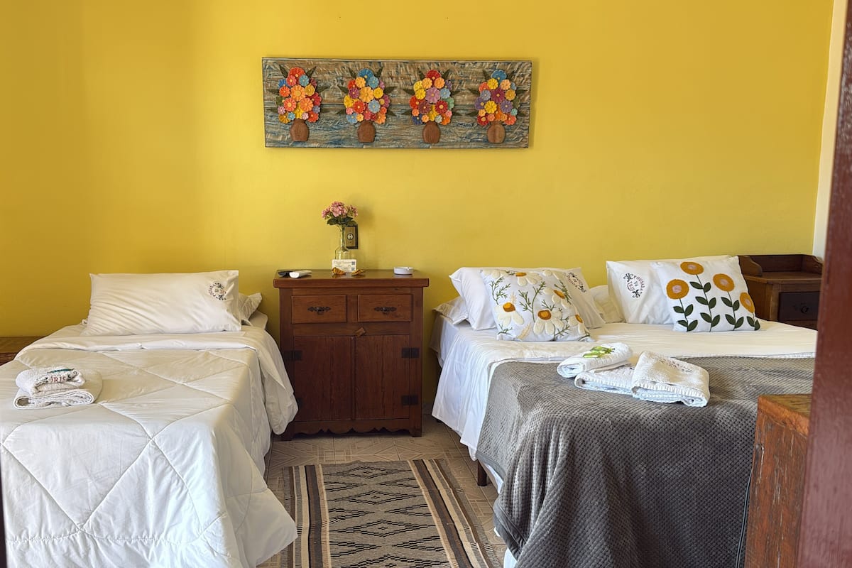 A room with two beds, a wooden nightstand with a vase of flowers, and a painting of flowers on the wall.