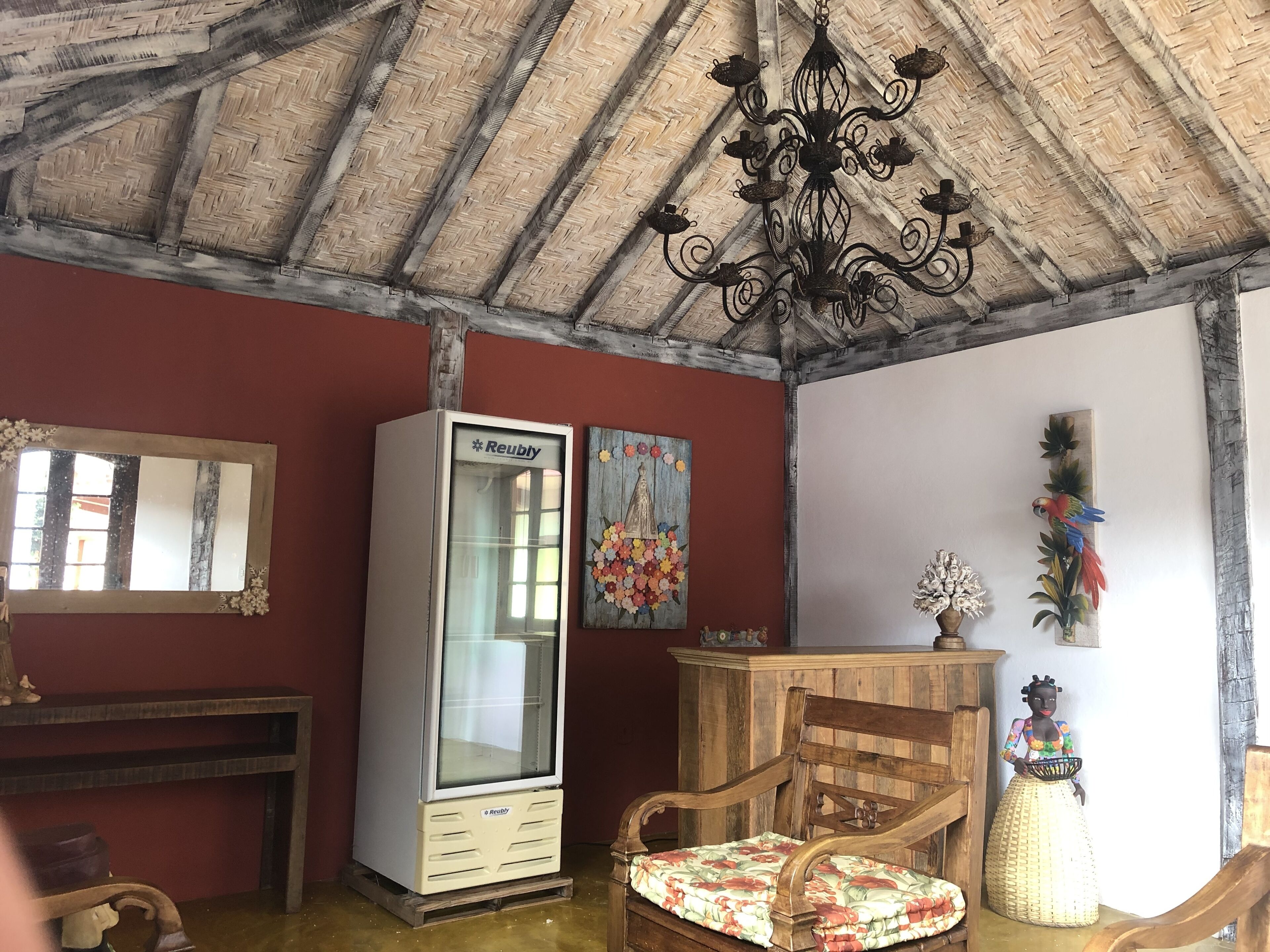 Een kamer met een houten plafond, een witte koelkast, een houten stoel met een bloemenkussen, een houten kast en een spiegel aan de muur.