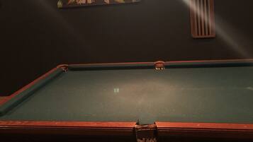 A pool table with a green felt surface, a wooden border, and a corner pocket. There is a wall-mounted light fixture above the table, and a framed floral artwork is visible on the wall.