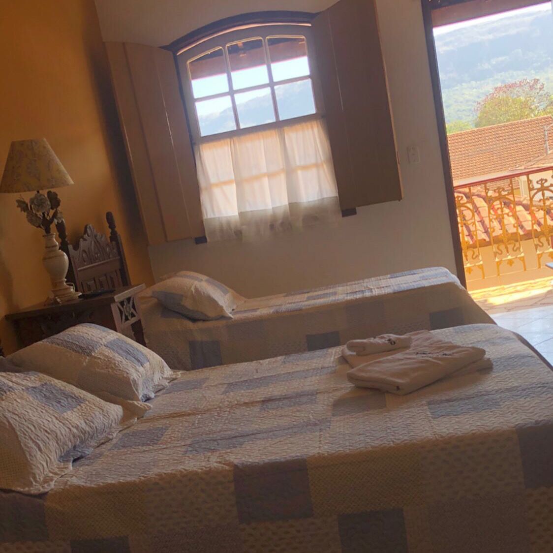 Habitació amb dos llits, una finestra amb cortines, una tauleta de nit amb làmpada i vista a un balcó.
