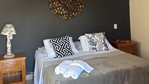 Un dormitorio con cama, mesita de noche de madera, lámpara, un adorno en la pared y una lámpara colgante.