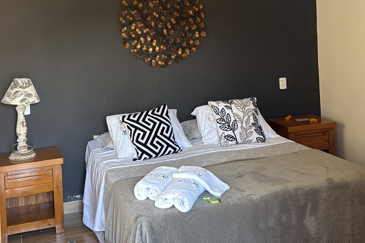 A bedroom with a bed, a wooden nightstand, a lamp, a decorative wall piece, and a hanging light fixture.