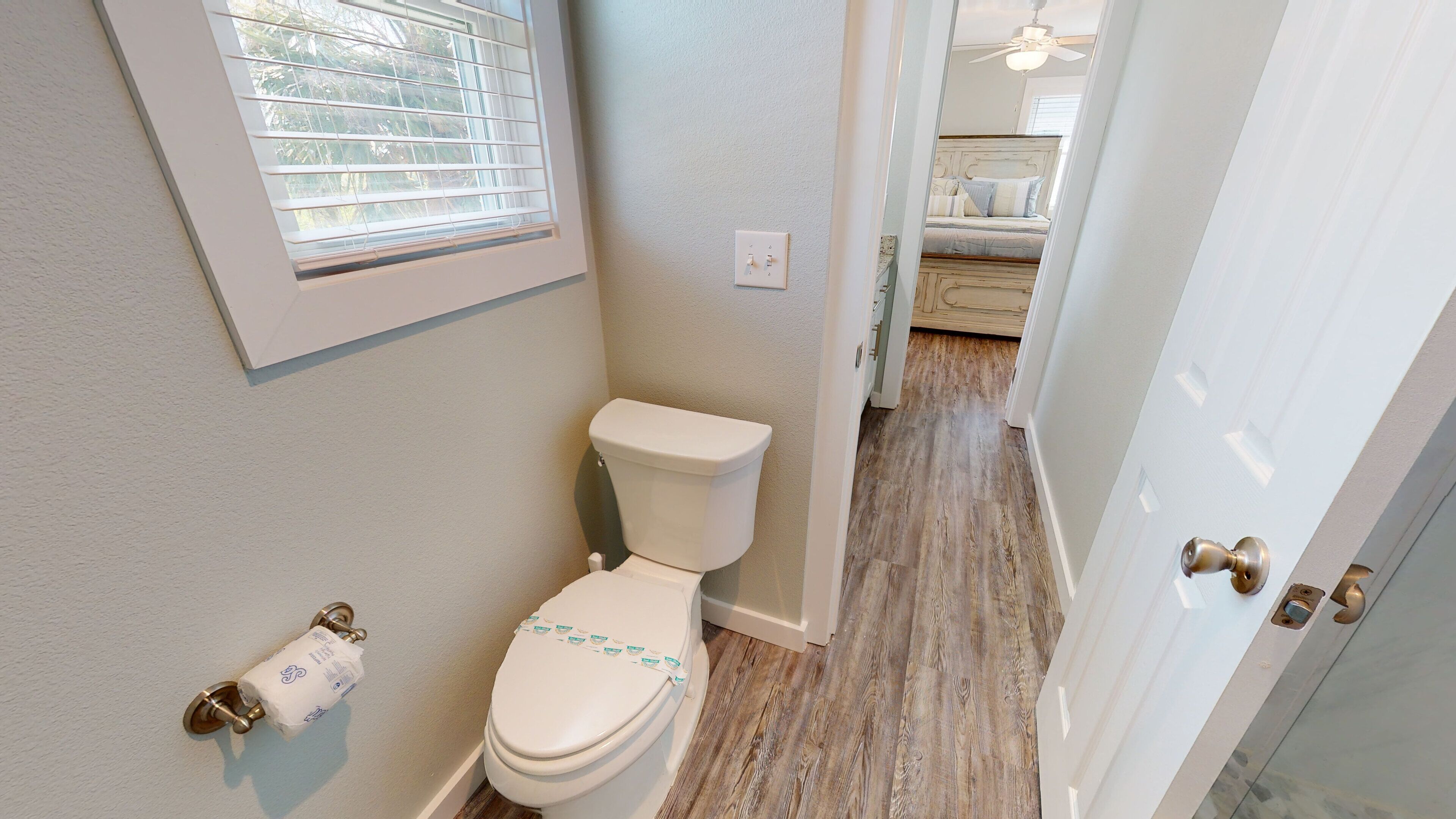 A bathroom with a toilet, a window with blinds, and a door leading to a bedroom.