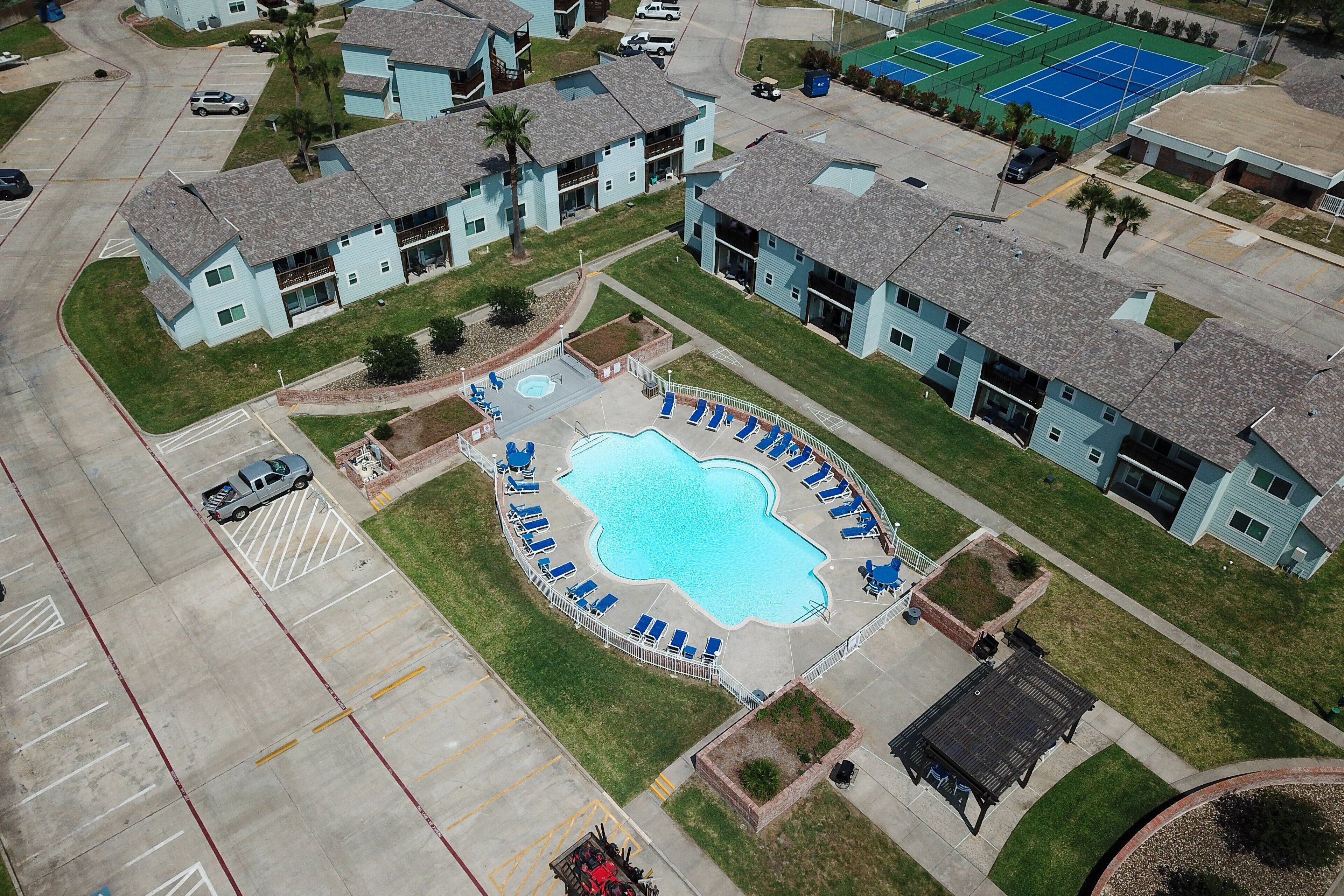 An aerial view of a residential area with a swimming pool surrounded by lounge chairs, a tennis court, and a parking lot.