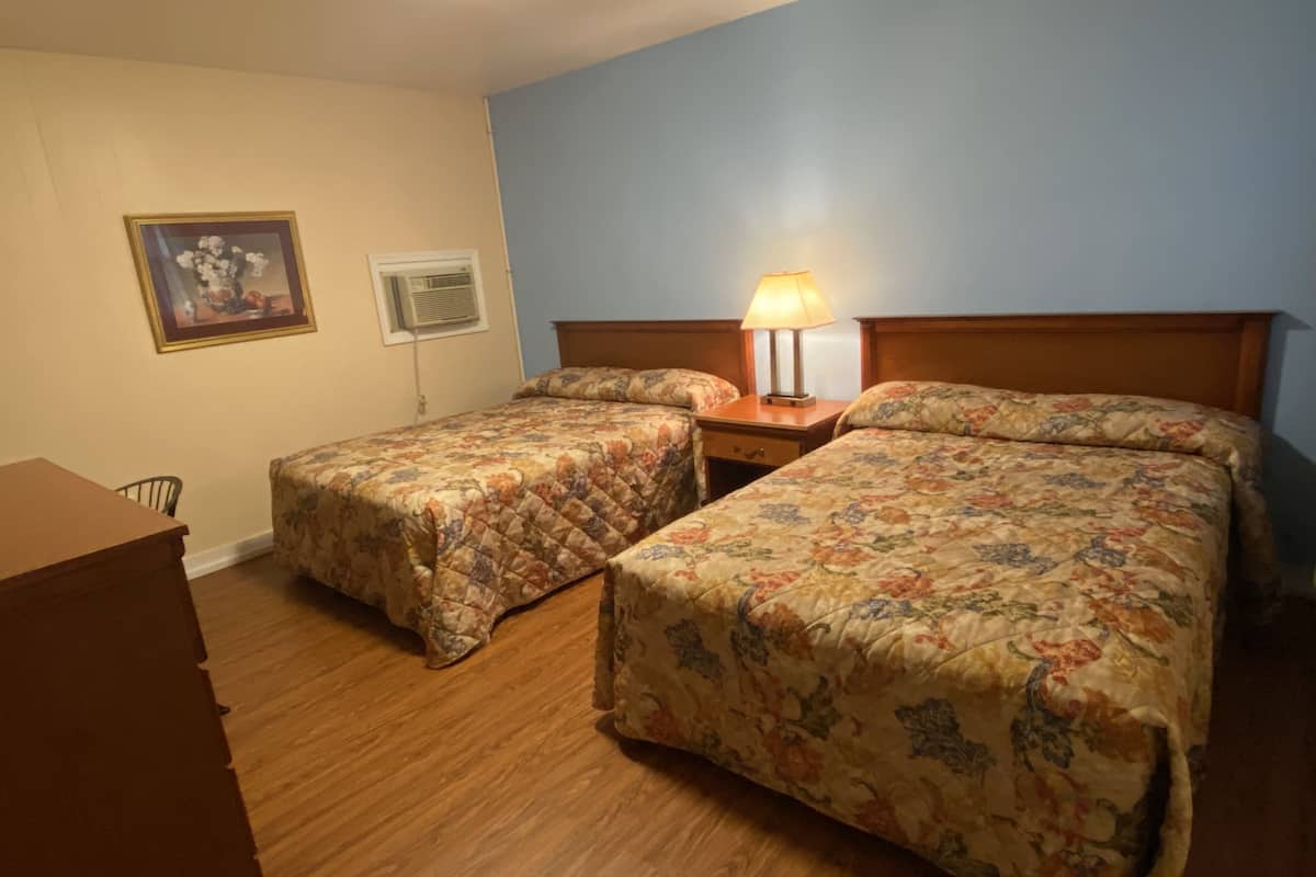 A hotel room with two single beds, a wooden dresser, a desk, a chair, a lamp, and a framed picture on the wall.