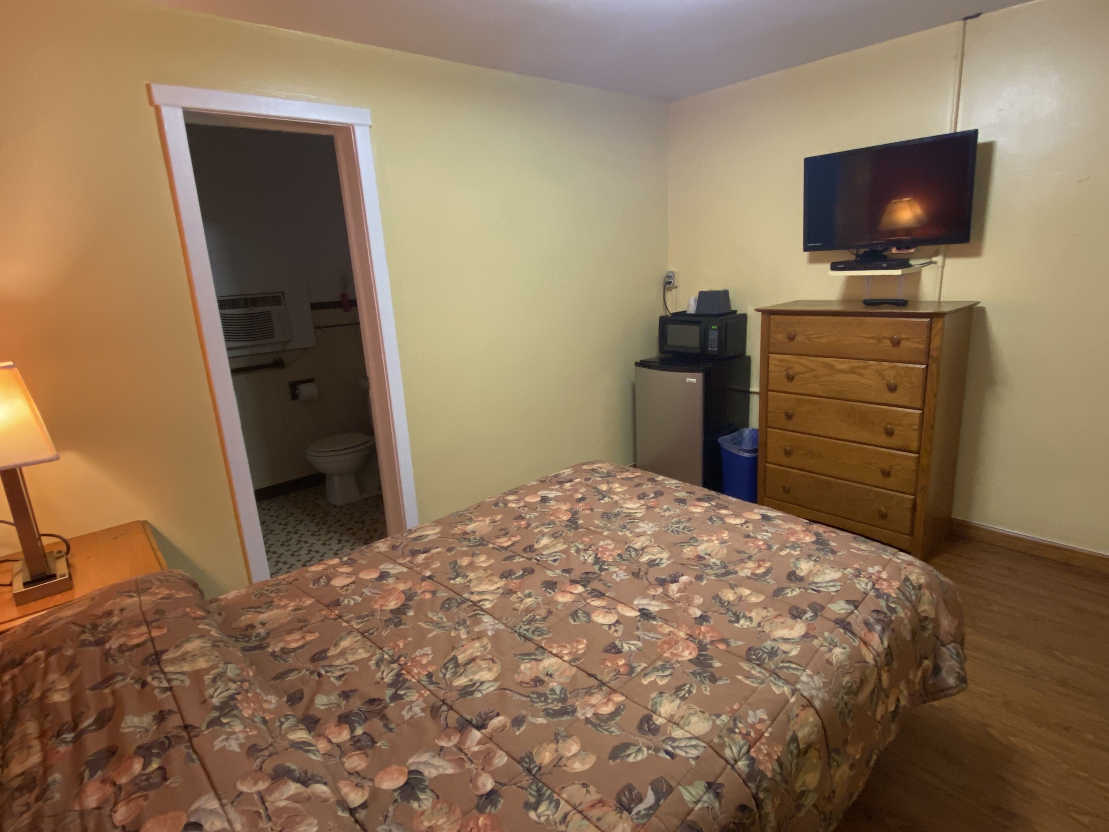 A bedroom with a bed, dresser, television, and a small kitchenette area.
