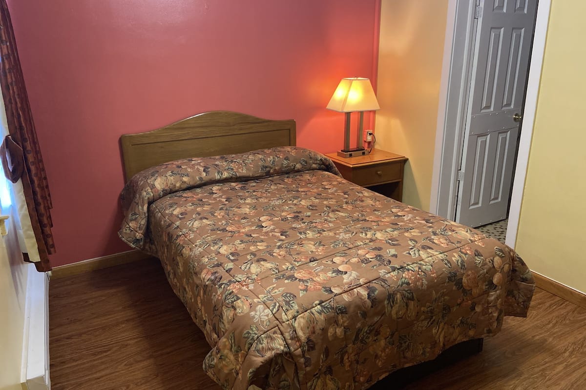 A neatly made bed with a floral bedspread, a wooden headboard, a bedside table with a lamp, and a door in the background.