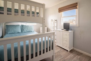 A room with a bunk bed, a desk with a computer, a window with blinds, and a view of buildings outside.