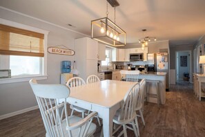 Une cuisine avec des meubles blancs, une table à manger avec des chaises, un luminaire suspendu et un panneau indiquant « MAISON DE PLAGE ».