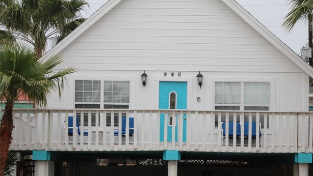 A white building with a turquoise door and windows, a balcony with blue chairs, and a palm tree in the foreground. - SUPER LOCATION, MIDDLE OF THE ISLAND (South Padre Island)