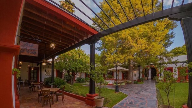An outdoor patio area with a covered seating area, potted plants, and a pathway leading to a building entrance. - Hotel Hostal El Farol (Parras)