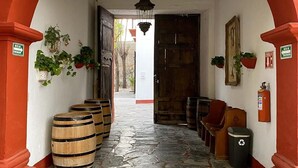 A corridor with a stone floor, wooden barrels, potted plants, and a wooden door. - Hotel Hostal El Farol (Parras)