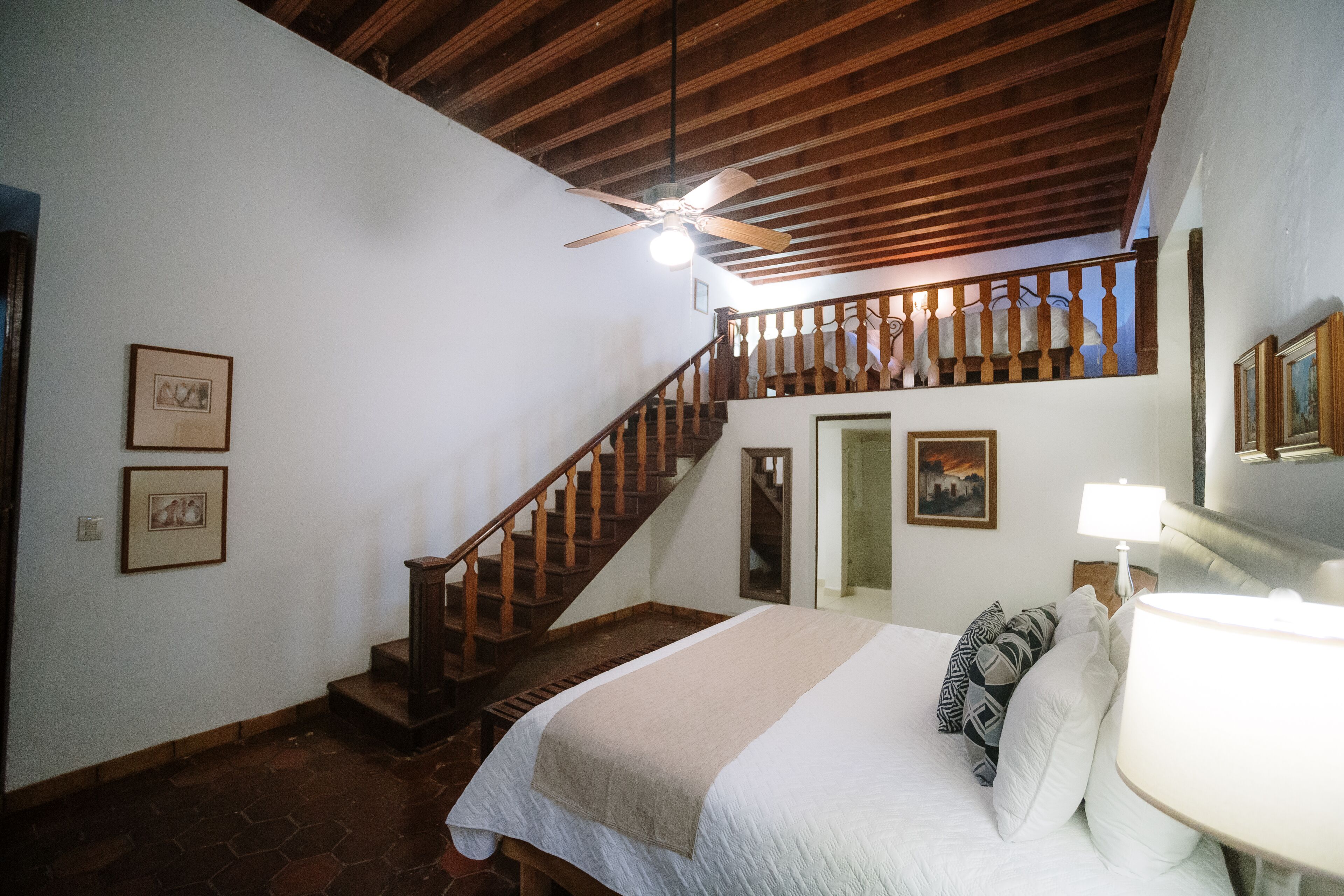 A bedroom with a bed, a nightstand, a lamp, a ceiling fan, a staircase, and framed pictures on the walls.