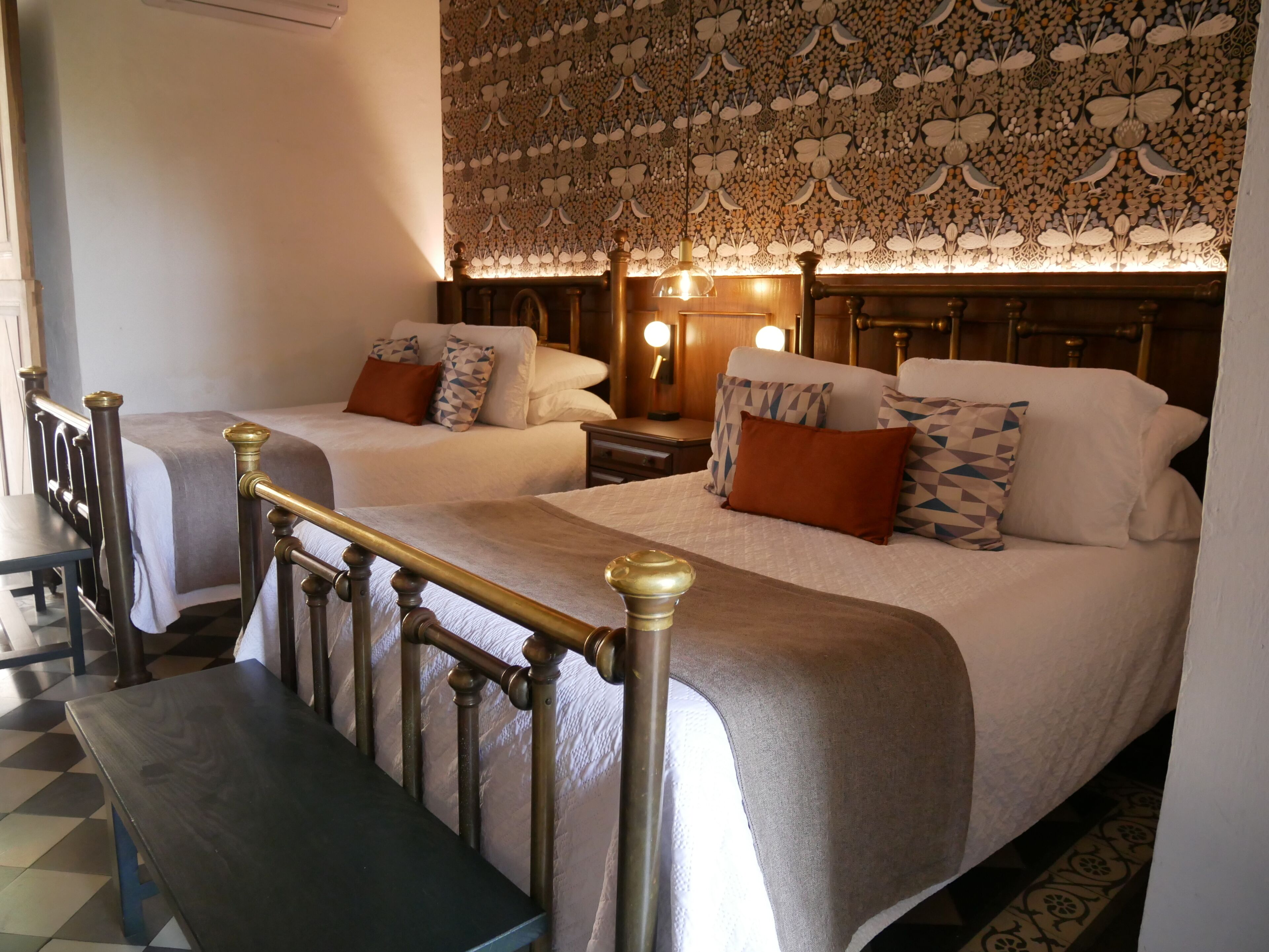 A hotel room with two beds, a bench, a patterned ceiling, and a nightstand with a lamp.