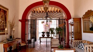A spacious hallway with a tiled floor, a wooden ceiling, and a red archway leading to a balcony. There are potted plants, a chair, and a sofa along the walls. - Hotel Hostal El Farol (Parras)