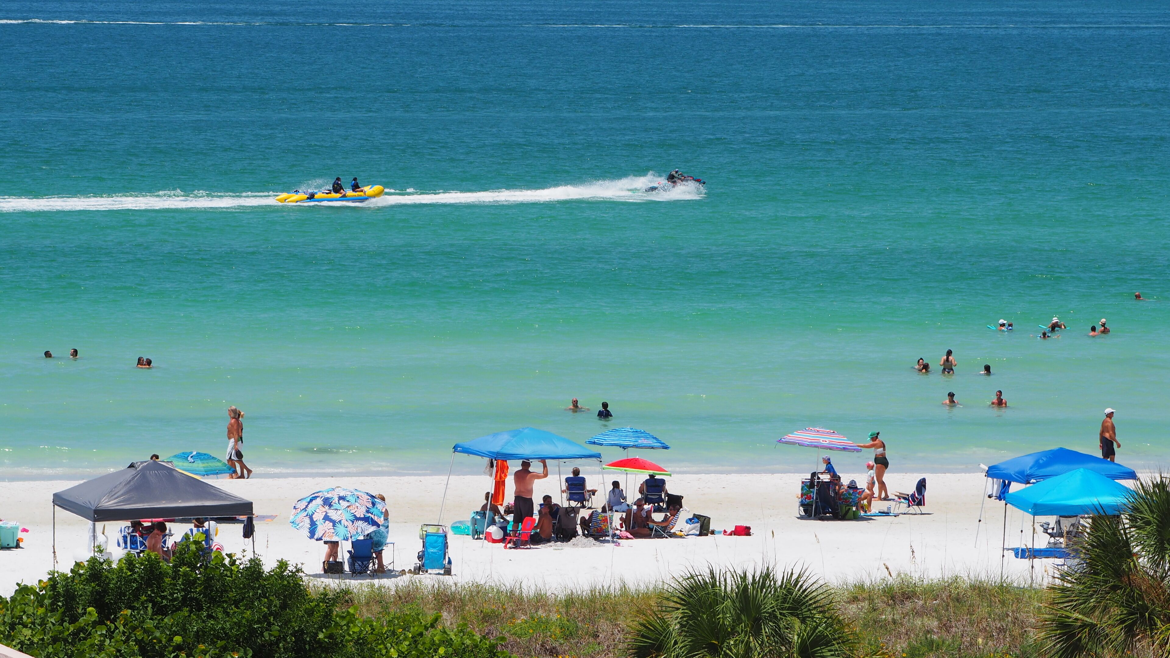 A beach with people swimming and a jet ski in the water. There are umbrellas and beach chairs on the sand.