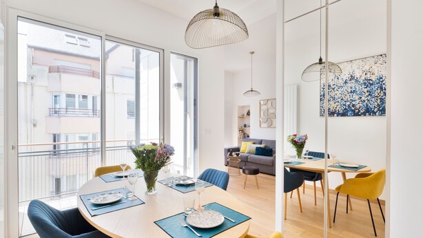 Un espace repas moderne avec une table ronde, des chaises bleues et une chaise jaune. Un balcon offre une vue sur les bâtiments.