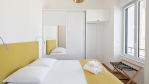 Une chambre d’hôtel avec un lit, une tête de lit jaune, un banc en bois, une fenêtre et une armoire.