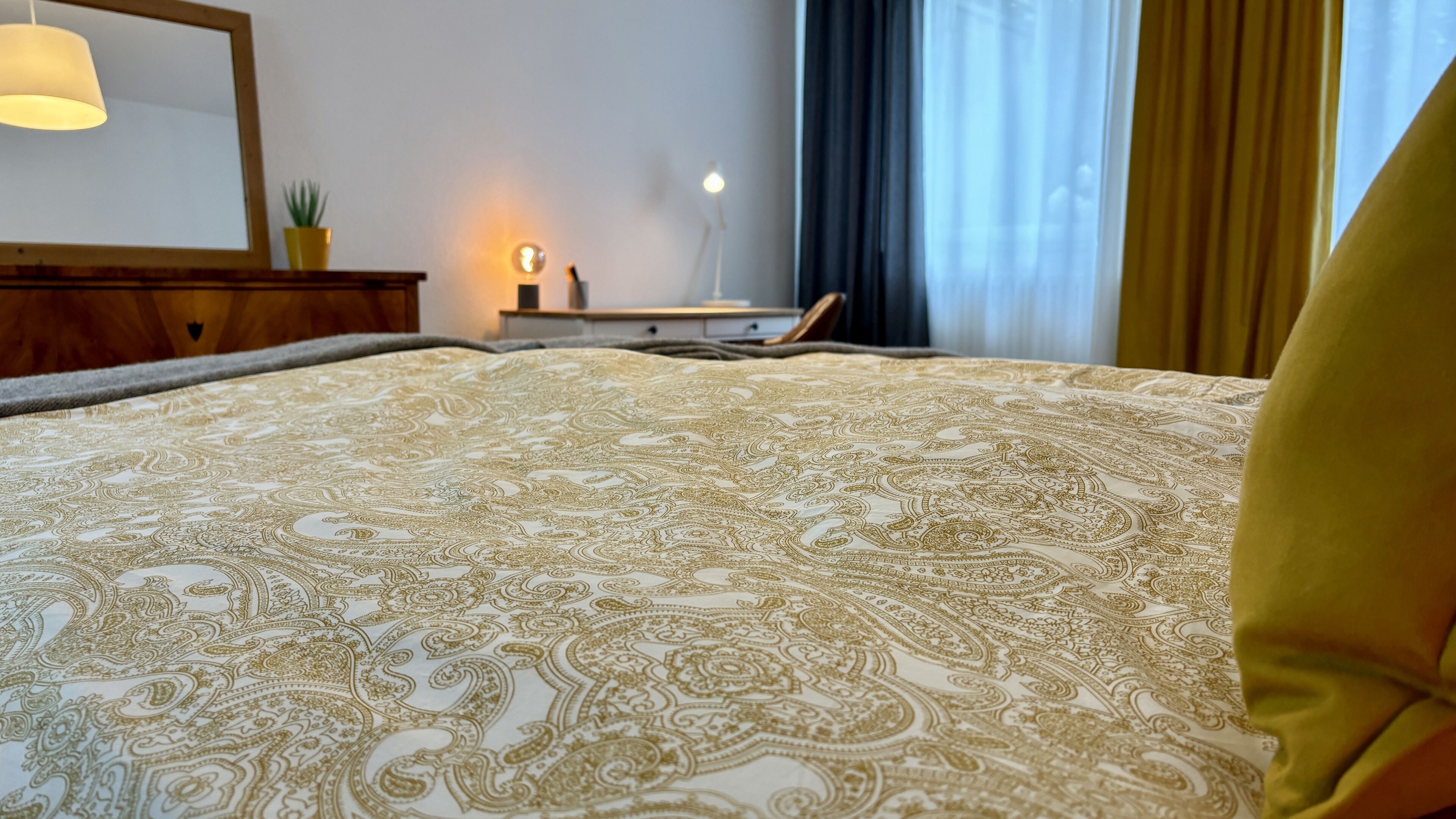 A neatly made bed with a patterned bedspread, a bedside lamp, and a nightstand with a lamp and a plant.