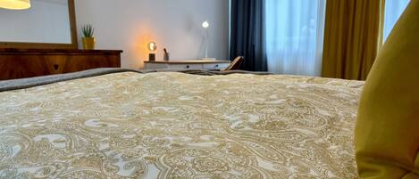 A neatly made bed with a patterned bedspread, a bedside lamp, and a nightstand with a lamp and a plant.