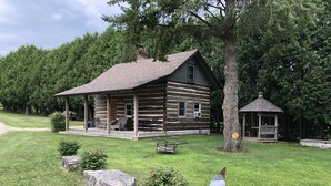 Une cabane en rondins avec un porche, un petit kiosque et un grand arbre dans une zone herbeuse.