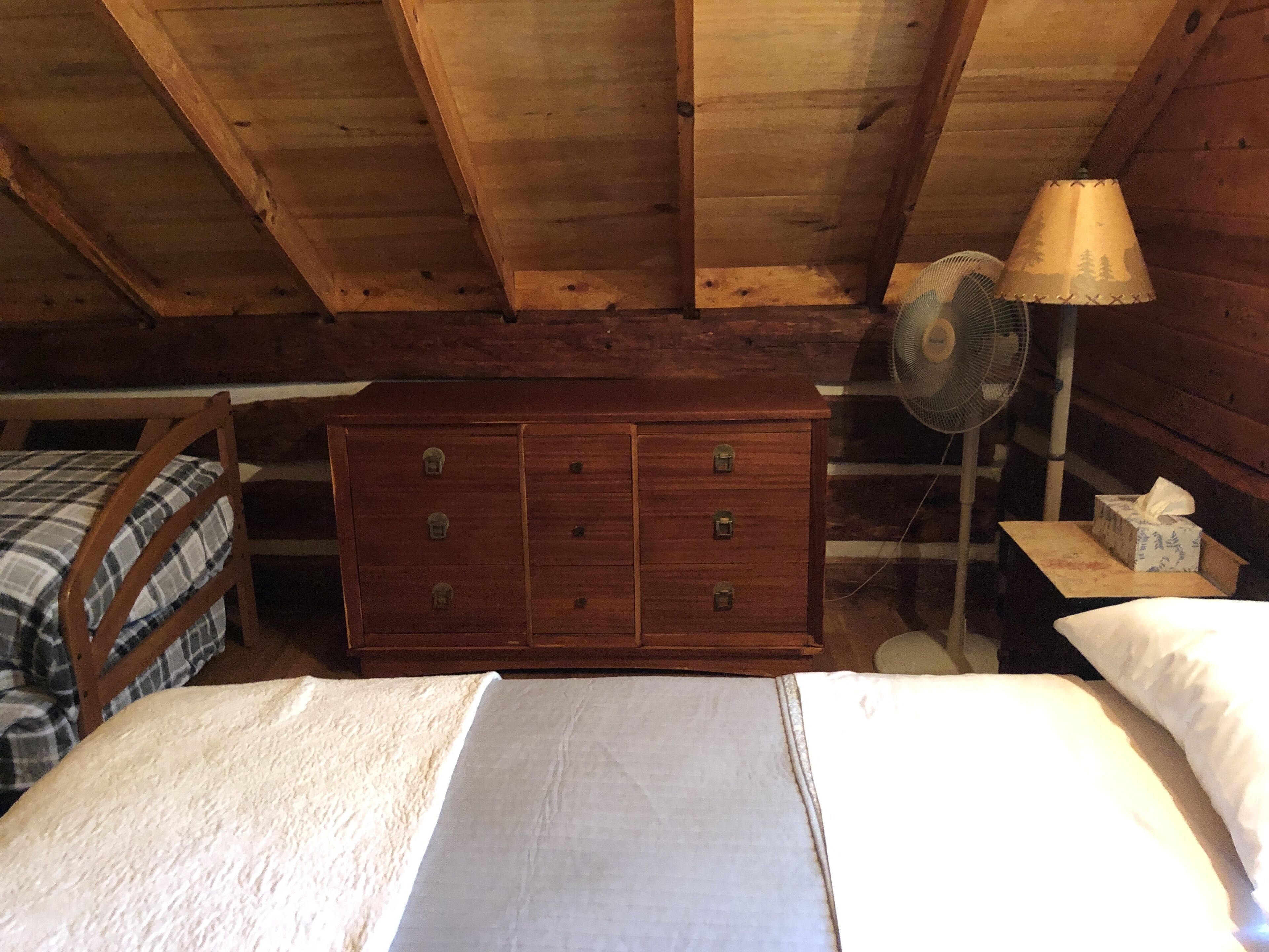 Ein Zimmer in einer Holzhütte mit einem Bett, einem Schminktisch und einem Ventilator.