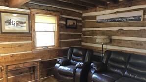 L'intérieur d'une cabane en rondins, avec un bureau en bois, des fauteuils inclinables en cuir, une fenêtre avec des stores et des tableaux encadrés accrochés aux murs.