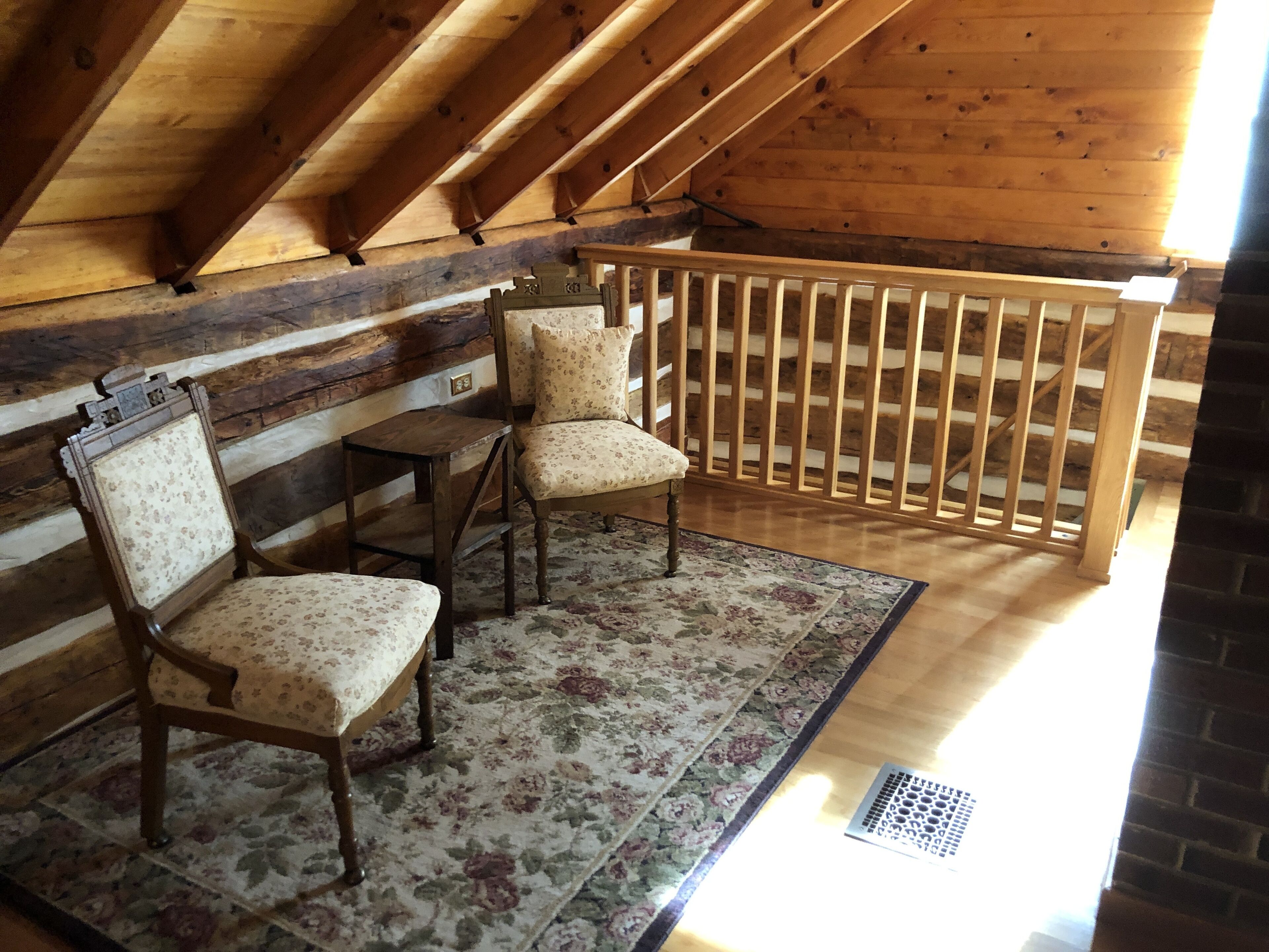 Une pièce mansardée chaleureuse avec des poutres apparentes, une rambarde en bois et deux fauteuils rembourrés recouverts d’un tissu à motifs floraux.