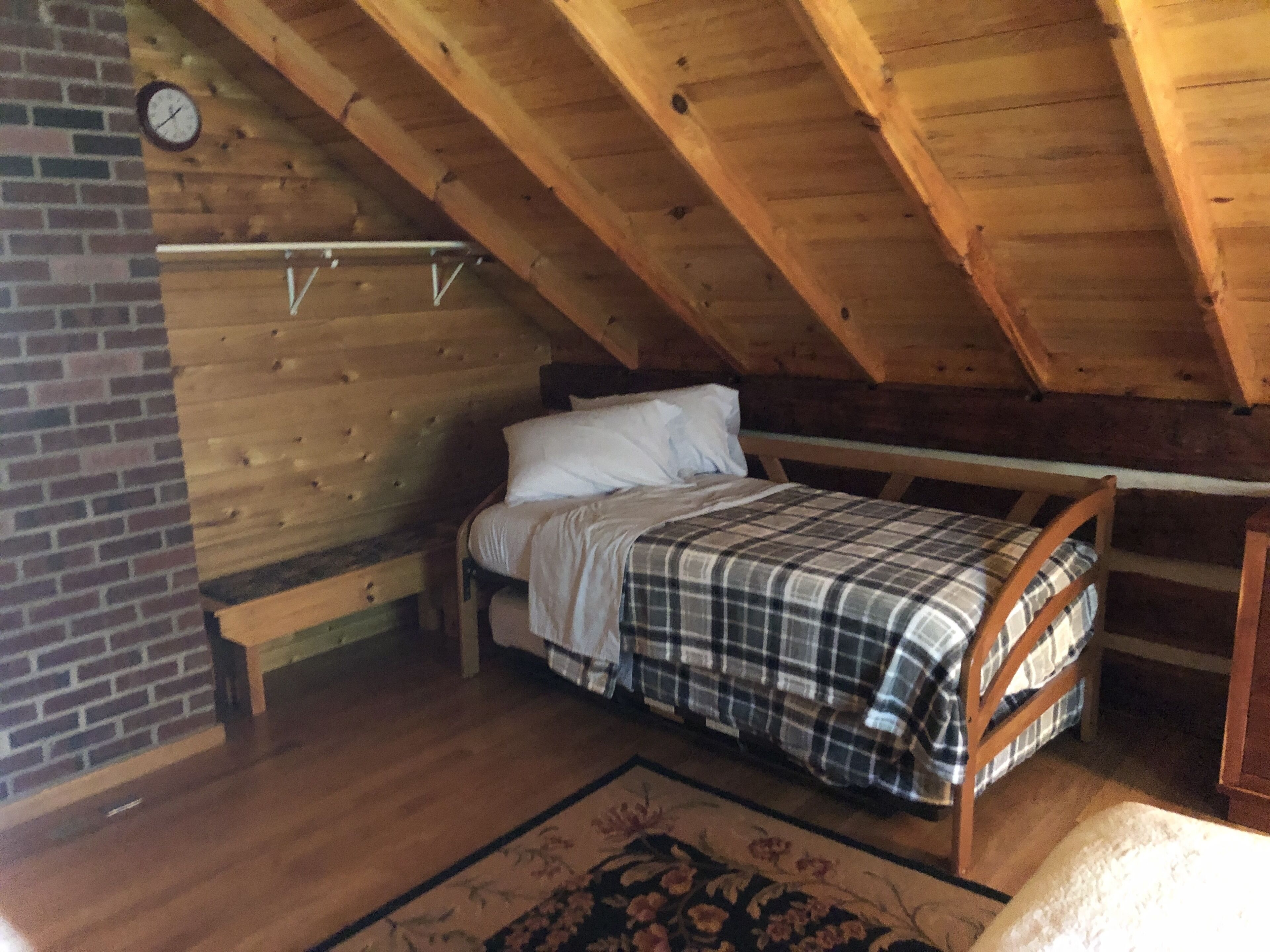Ein Schlafzimmer im Loft-Stil mit Holzbalkendecke, Backsteinwand, einem Bett mit karierter Decke, einer Bank und einer Uhr.
