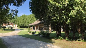 Un chalet rustique en rondins de bois, doté d’un porche et de meubles d’extérieur, entouré d’une végétation luxuriante et accessible par une allée de gravier.