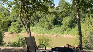 A campsite with a fire pit and two chairs facing a river, surrounded by trees. - Aggie Game day in the country outside of College Station!  (Cameron)