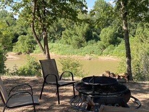 Een camping met een vuurkuil en twee stoelen gericht op een rivier, omringd door bomen.