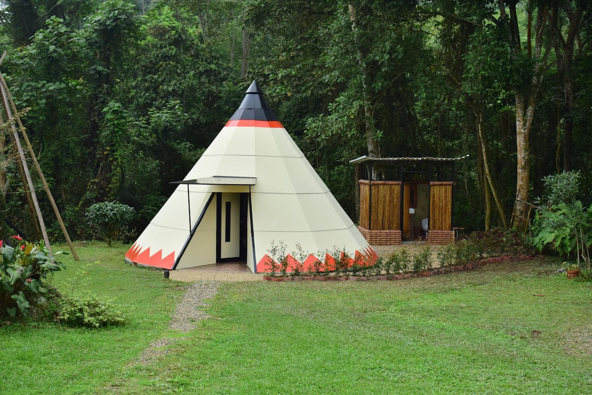 A conical tent with a door, surrounded by a garden with red flowers and a small wooden structure.