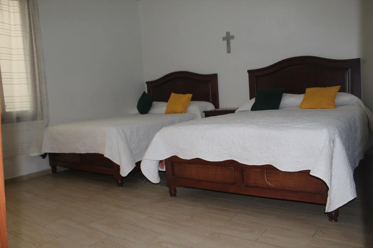 Deux lits en bois avec du linge de lit et des oreillers blancs, un plancher en bois et un crucifix fixé au mur.