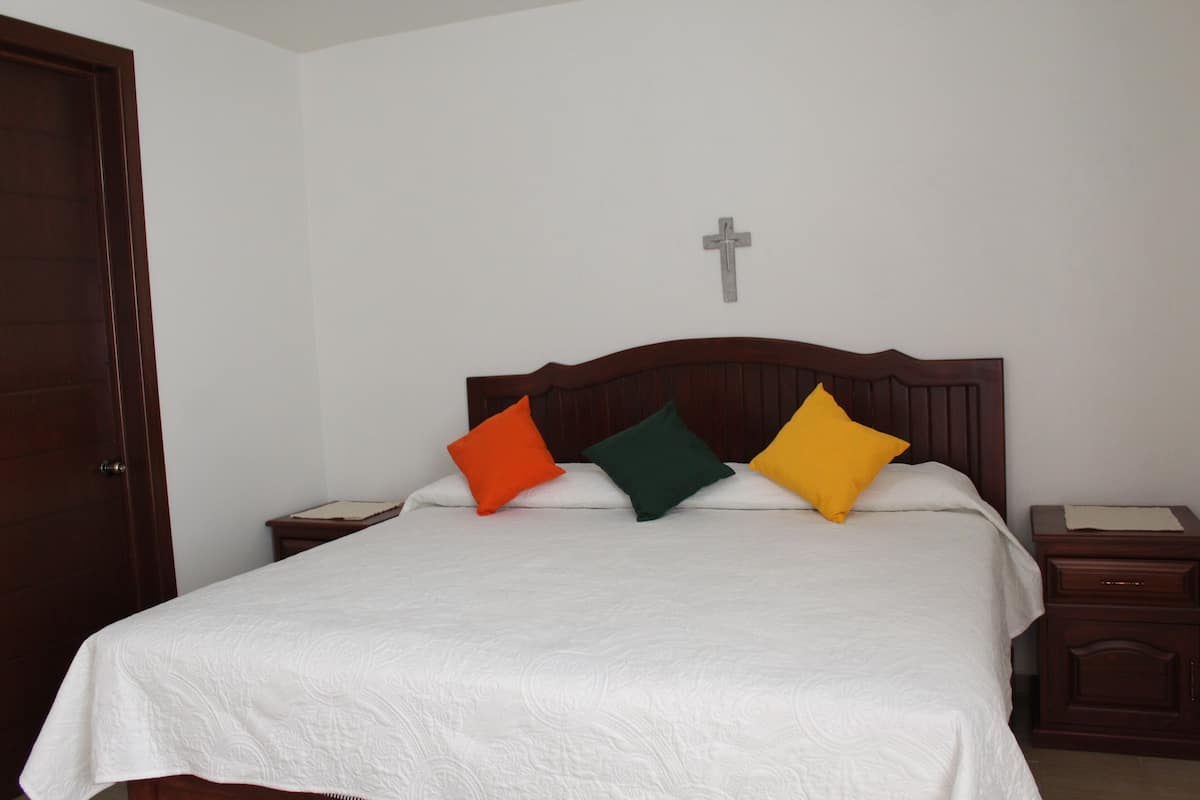 Un lit bien fait, recouvert d’une couvre-lit blanche et agrémenté de trois oreillers aux couleurs orange, verte et jaune. On y voit une tête de lit en bois, une petite table de chevet en bois avec une lampe, et une croix accrochée au mur au-dessus du lit.