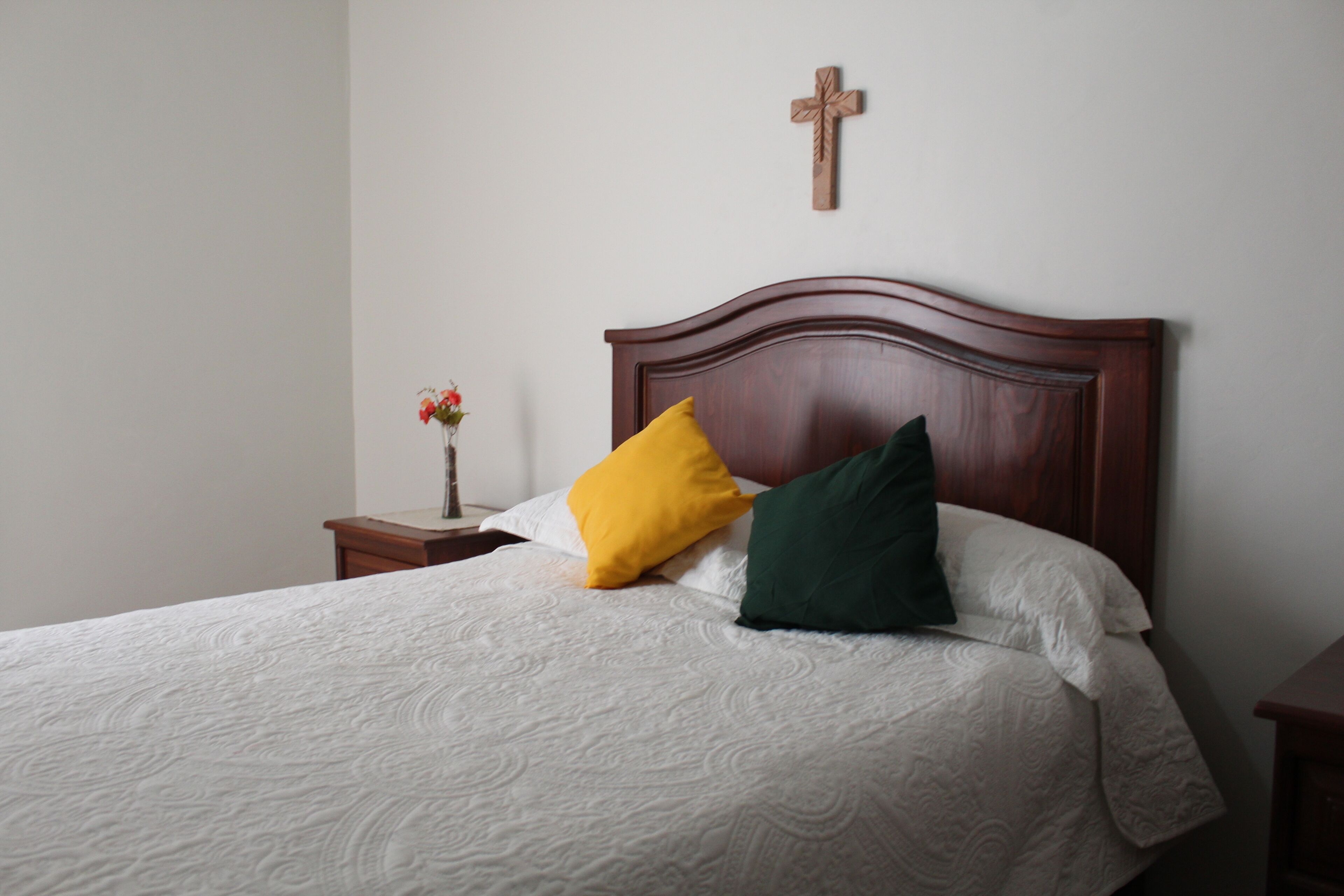 A neatly made bed with a white quilt and two pillows, a wooden cross on the wall, and a small bedside table with a vase of flowers.