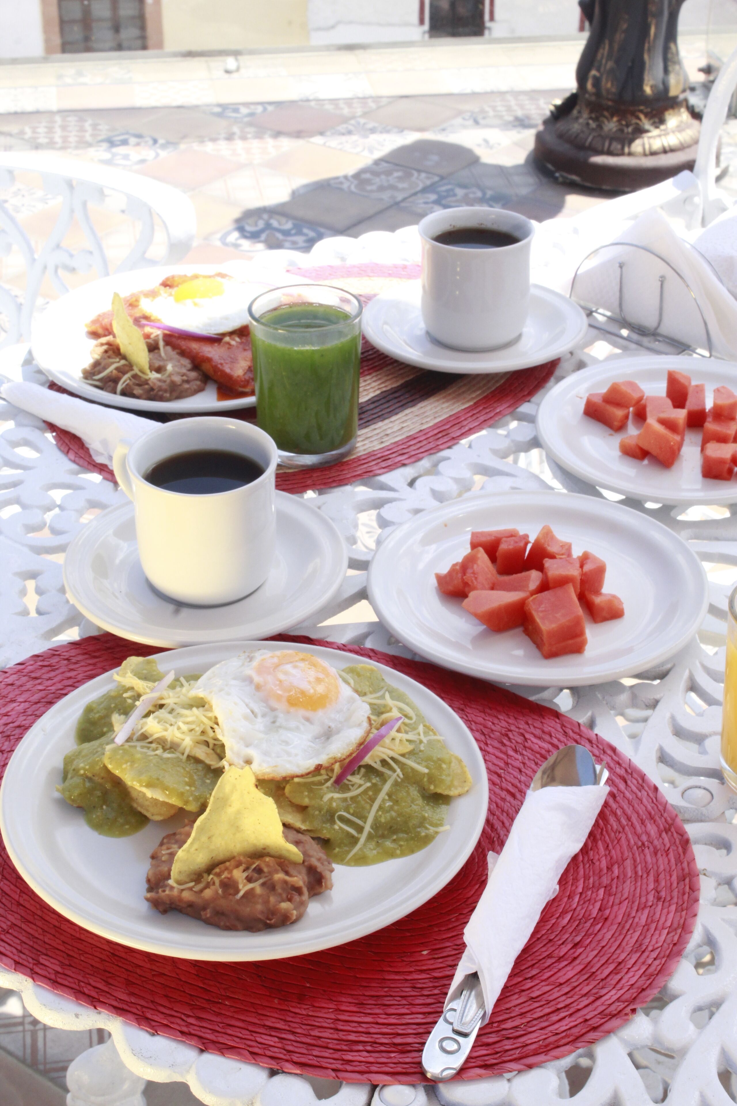 Uma mesa posta com diversos pratos, incluindo um com feijão e ovo frito, outro com cubos de melancia, uma xícara de café e uma bebida verde.