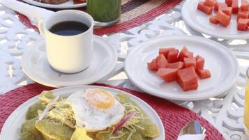 Uma mesa posta com diversos pratos, incluindo um com feijão e ovo frito, outro com cubos de melancia, uma xícara de café e uma bebida verde.
