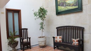 A room with a tiled floor, a wooden bench with floral cushions, a potted plant, a chair, and a framed painting on the wall.
