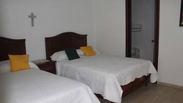 A hotel room with two single beds, a wooden headboard, a white cross on the wall, and a door leading to a bathroom.