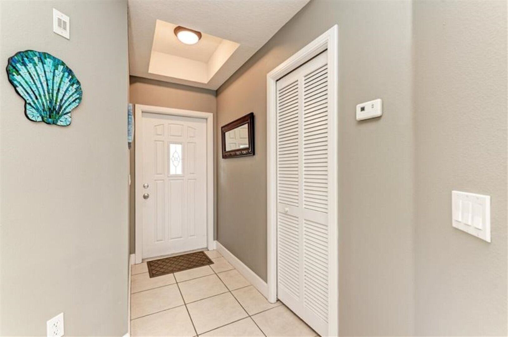 Un pasillo con una puerta blanca, un armario blanco con puertas persianas y una pequeña concha decorativa en la pared, además de una luz empotrada en el techo.