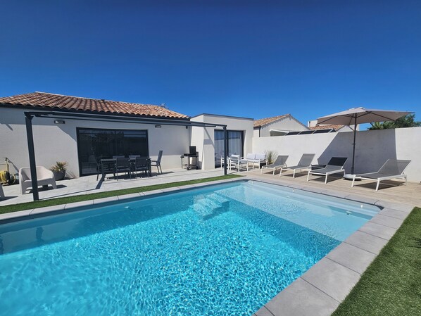 A modern poolside area with a clear blue pool, lounge chairs, and a patio with a table and chairs.