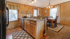 A kitchen with wooden cabinets, a granite countertop, a sink, and a dining area with a table and chairs. -  Large-Group Cabin near Luray · Sleeps 12 · Hot Tub & Mountain Views (Rileyville)