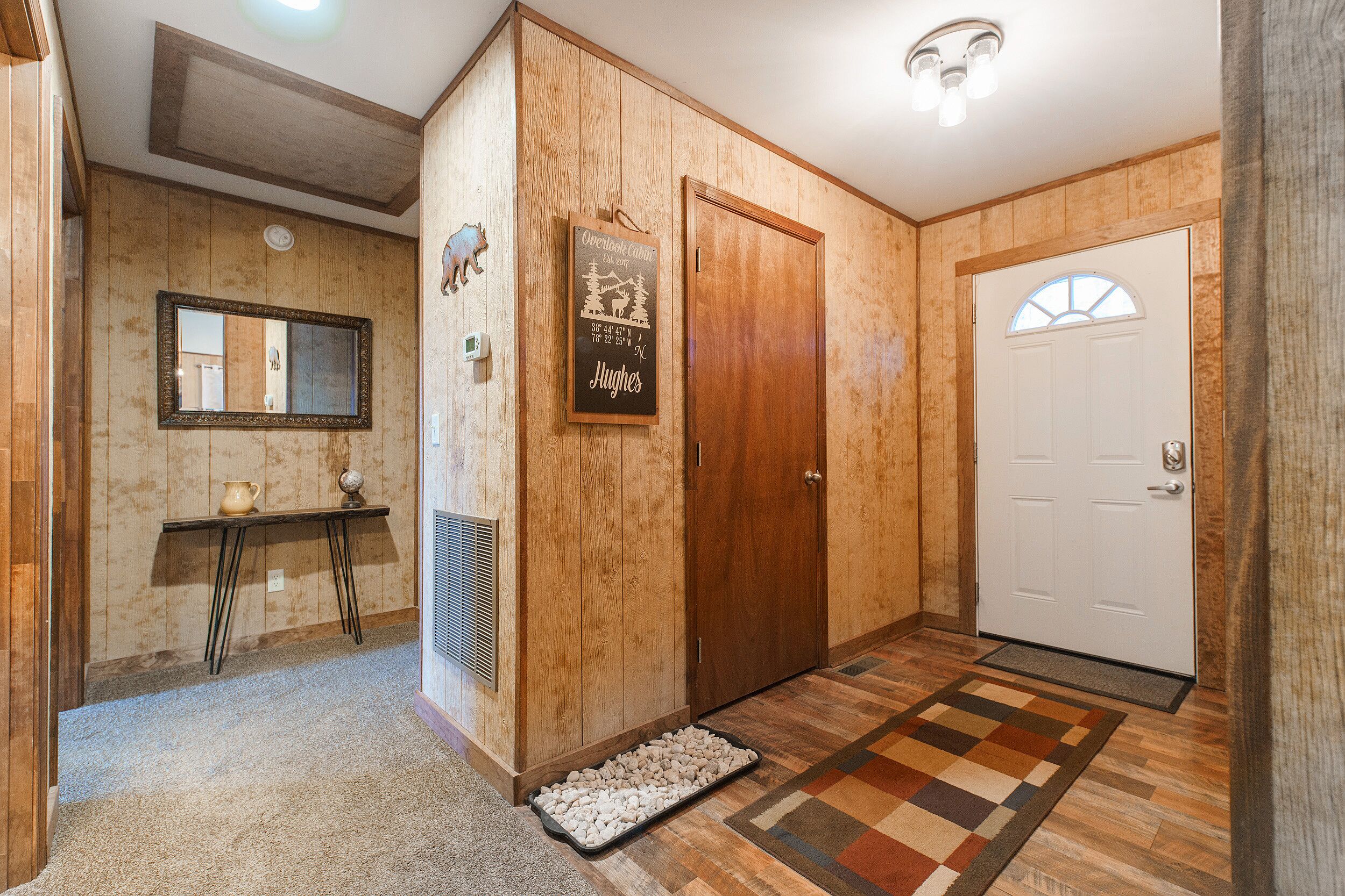 A wooden-paneled hallway with a mirror, a welcome sign, a door, and a patterned rug.