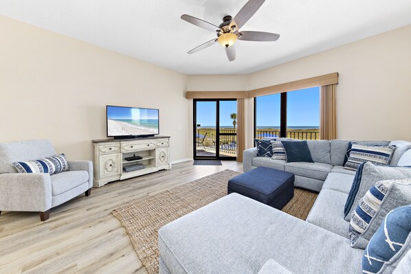 A modern living room with a large sectional sofa, a flat-screen TV, a ceiling fan, and a view of the beach through large windows. - Phoenix VI 111 | Oceanfront | Private Balcony | Resort Amenities | My Beach Getaways (Orange Beach)