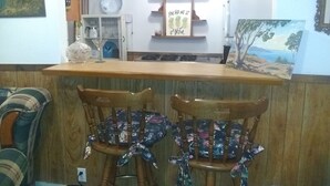 Een restaurantinterieur met een houten toonbank, twee houten stoelen met bloemenkussens, een bordje met de tekst 'ORDER HERE', en diverse wanddecoraties zoals een bord, een ingelijste foto en een plank met spullen.