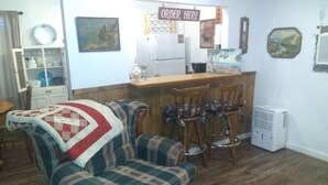 Een woonkamer met een geruite fauteuil, een eettafel met stoelen, een keuken met een bord waarop 'ORDER HERE' staat, en een wandairconditioner.